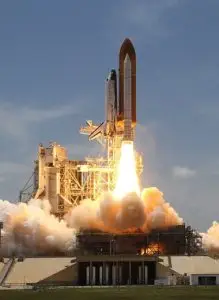 A space shuttle launches vertically, showing bright flames and smoke from the boosters against a clear sky. The launch pad infrastructure is visible beneath the shuttle.