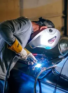 A person wearing gloves and a protective mask is welding metal, with bright sparks visible at the welding point.