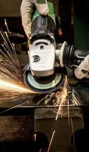 A gloved hand uses an angle grinder to smooth a piece of metal, producing sparks.