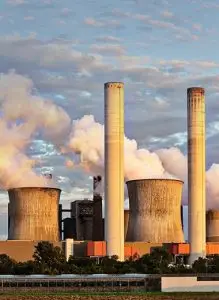 A large industrial power plant with four tall smokestacks emitting white smoke, set against a cloudy sky. There is greenery in the foreground.