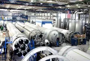 An industrial complex with several large rocket bodies in various stages of assembly, surrounded by scaffolding and machinery. An American flag and various equipment are visible in the background.