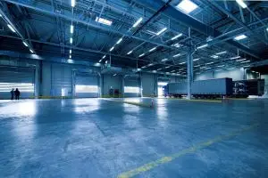 Spacious industrial warehouse with high ceilings, overhead lights, and multiple open loading docks. A large truck is parked at one dock, and three people stand in the distance.