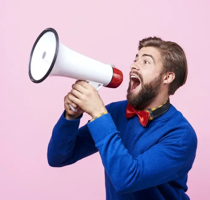 A bearded man shouting into a megaphone on a pink background, embracing Loud Budgeting.