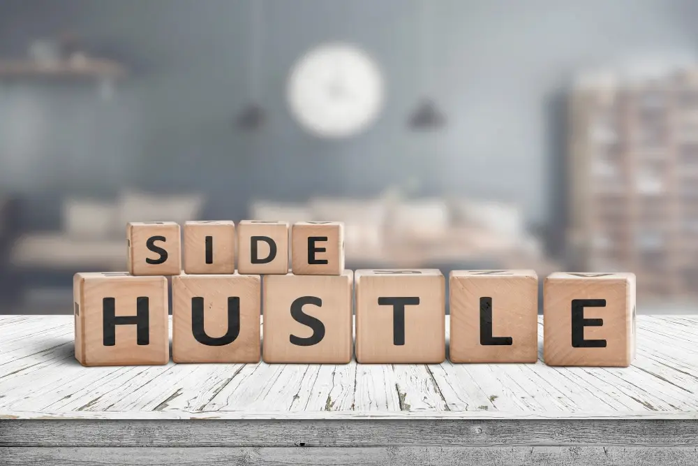 Wooden blocks on a table spelling out "Side Hustle" with a blurred interior background, symbolizing the balance between a 9 to 5 job and working a side hustle.