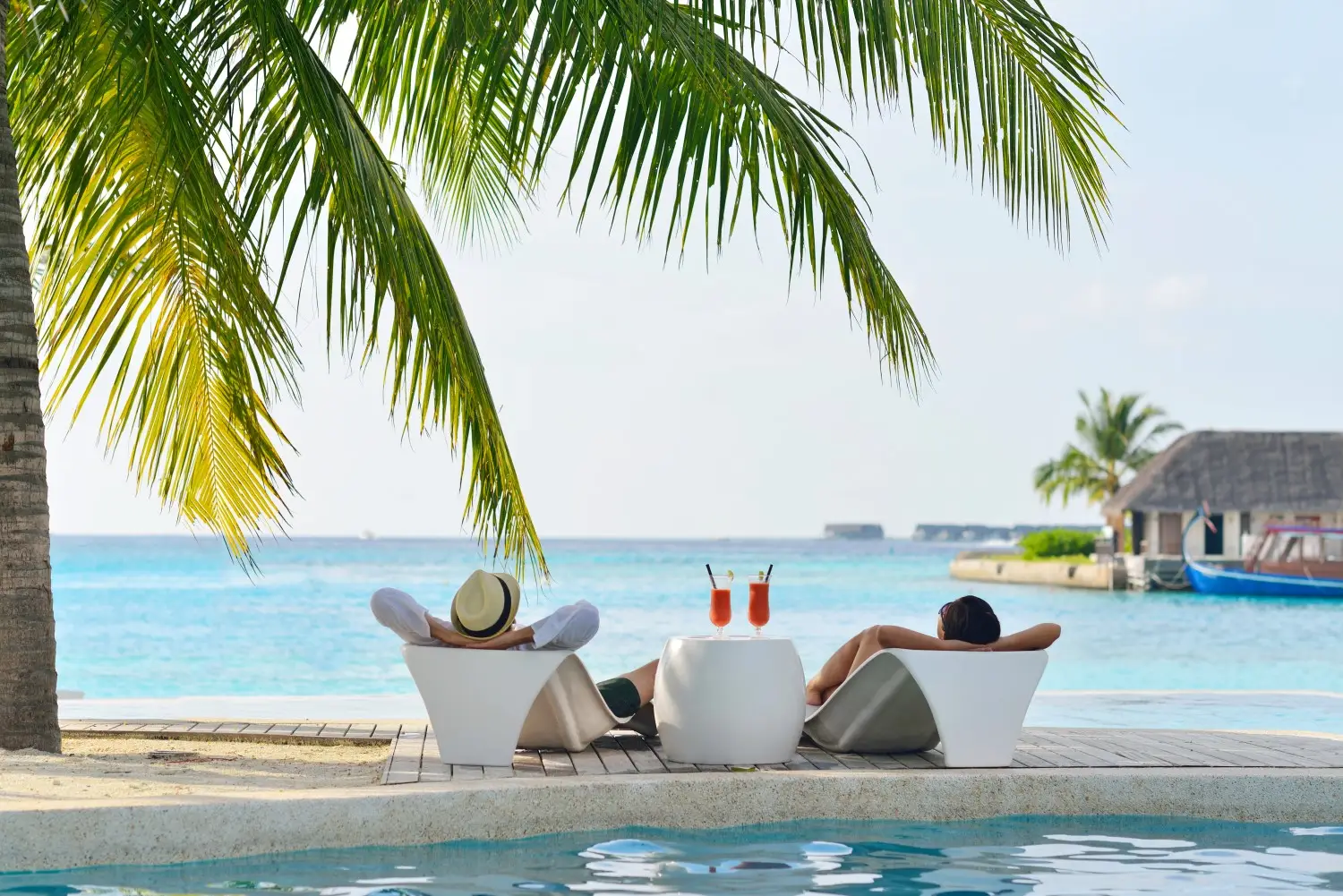 Two people relaxing in lounge chairs by a pool under palm trees, with a view of the ocean and a clear blue sky, enjoying their budget-friendly vacation.