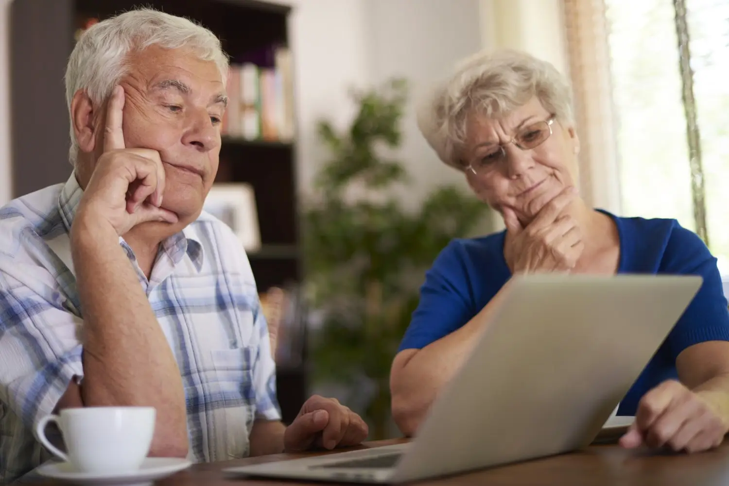 An elderly couple, a man and a woman, look intently at a laptop screen while sitting at a table with a cup of coffee, discussing their retirement finances.