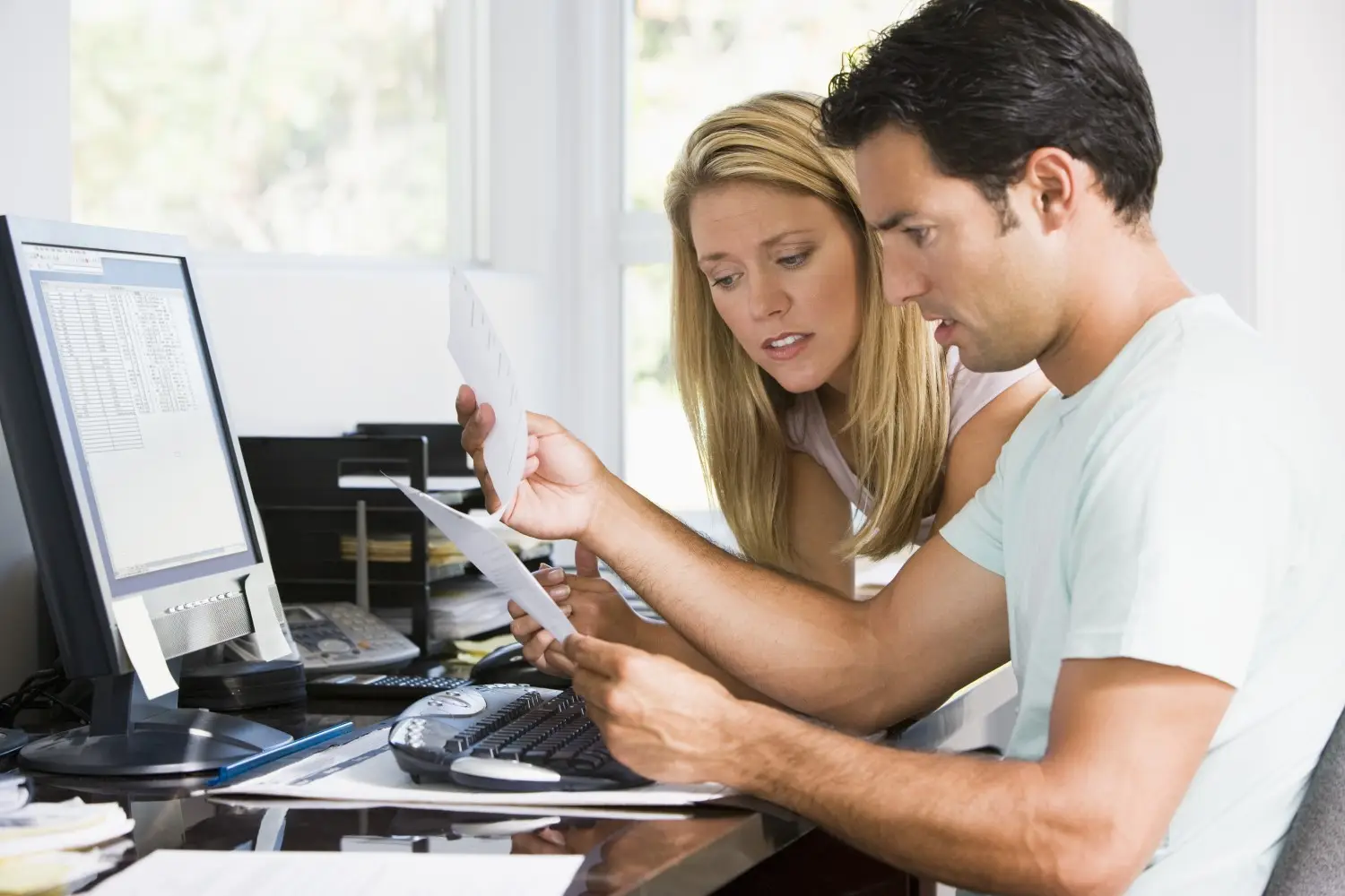 A man and woman are seated at a desk, looking at documents. A computer monitor displaying spreadsheets is on the desk along with various office supplies as they seek financial advice, showing concern that their payments exceed income.