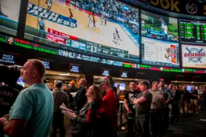 People watching multiple sports games and odds on large screens at a busy sports gambling venue.