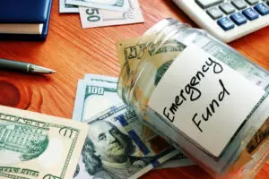 A glass jar labeled "foolproof emergency fund" filled with U.S. dollar bills, surrounded by more bills and a calculator on a wooden desk.