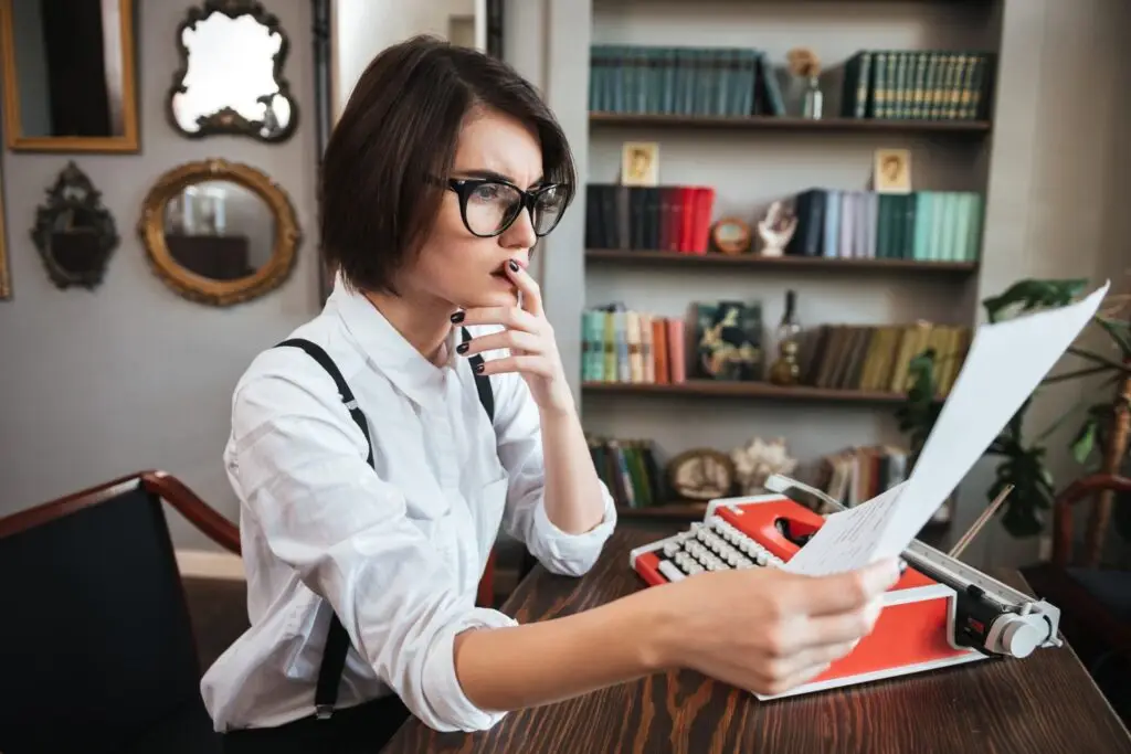 A person with short hair and glasses embraces the moment as they read a document while sitting at a desk with a vintage typewriter. Bookshelves and decorative mirrors surround them, reflecting their dedication to life growth. Side Hustles