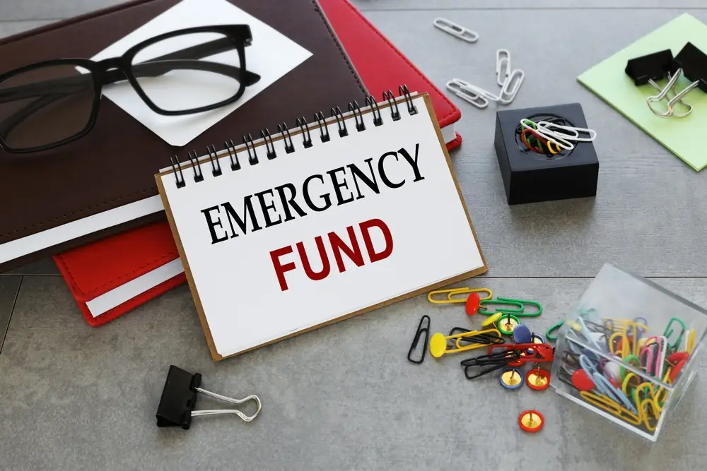 A notepad labeled "Emergency Fund" is displayed alongside eyeglasses, notebooks, and an assortment of paper clips on a desk, ready for you to build yours now.