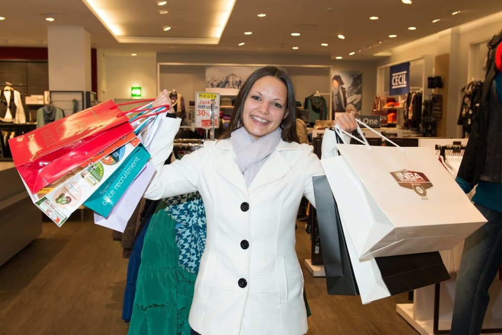 A person in a white coat smiles while holding multiple shopping bags in a clothing store, embracing the delights of impulsive spending. Various sale and recruitment signs are visible in the background, hinting at the excitement that costs money.