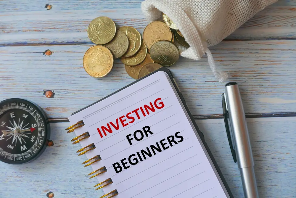 A notebook titled "Investing for Beginners 2024," a pen, a small bag with coins, and a compass on a wooden surface.