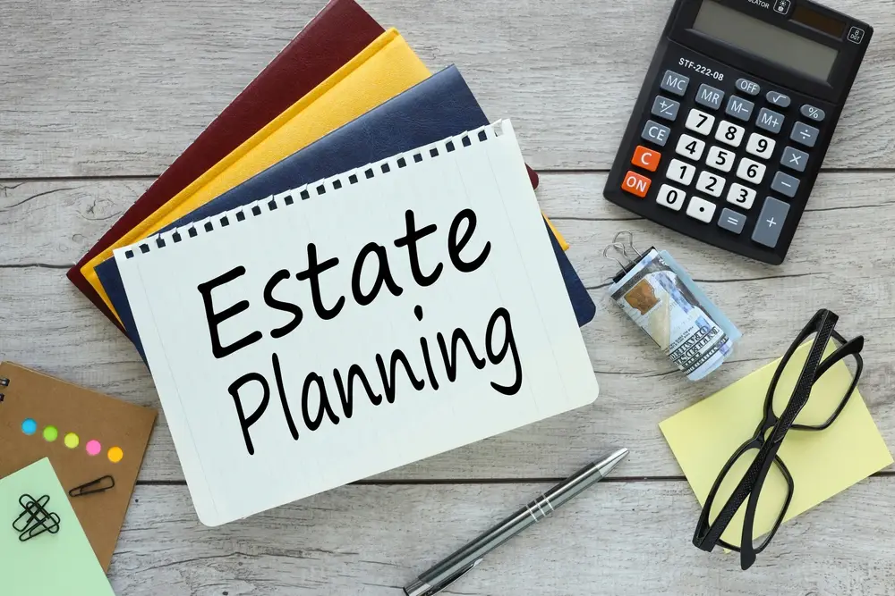 A notebook with "Estate Planning Guide" written on it is surrounded by colorful files, a calculator, glasses, sticky notes, a pen, and a miniature hourglass.