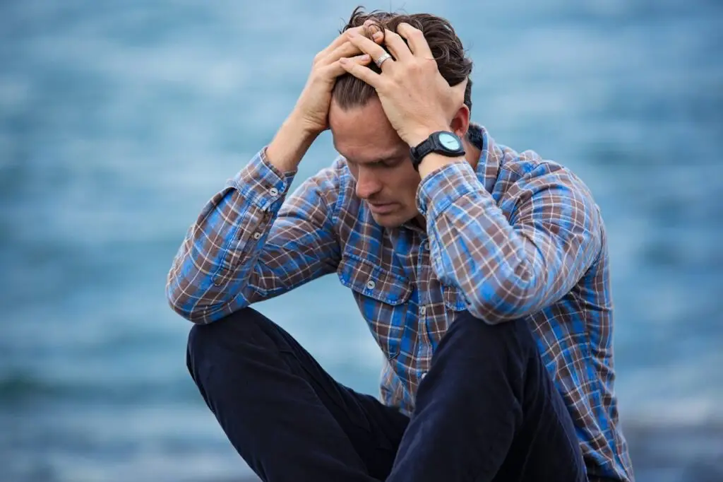 A man in a blue plaid shirt and dark pants sits by the water, holding his head with both hands.