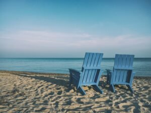 Two blue wooden chairs sit on a sandy beach facing calm blue ocean waters under a clear sky, offering the perfect spot to reflect on your retirement planning and secure your future.