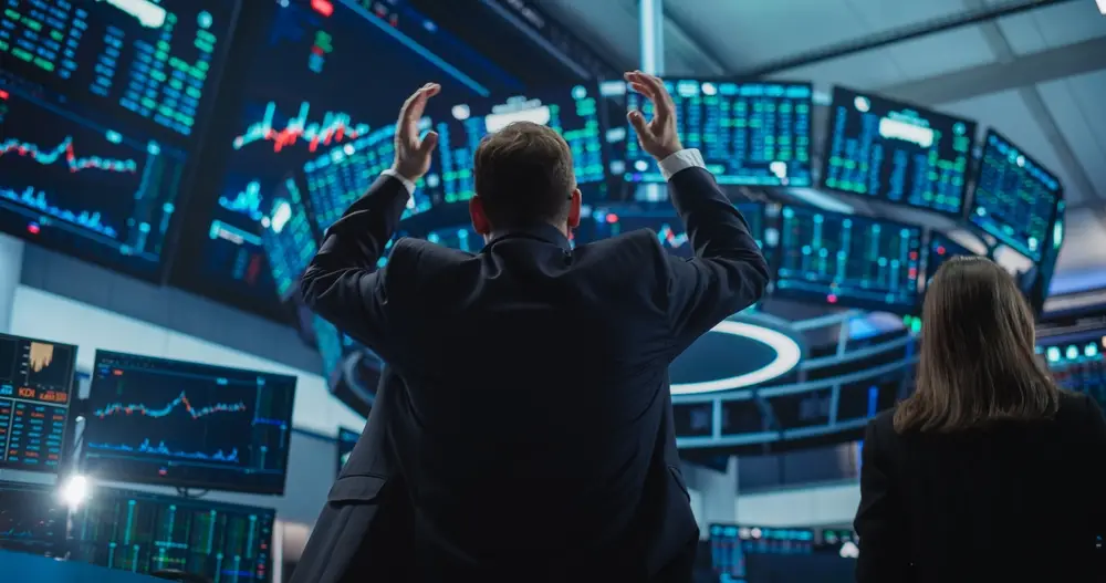 A person in a suit raises their hands while looking at multiple financial screens displaying stock market data showing a big drop; another person stands nearby facing the screens, concerned about your money.