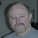 A middle-aged man with a light beard and mustache is wearing a gray shirt, looking at the camera. The background includes a dark object and candles, providing an intimate "About Me" feel.