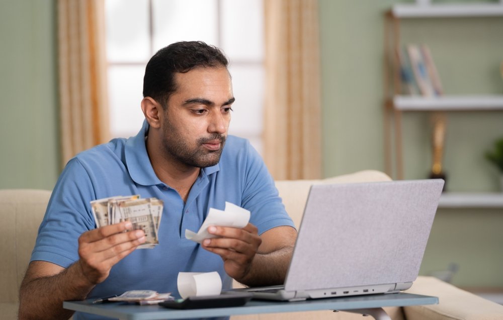 In uncertain times, a man in a blue shirt sits on a couch, intently focused on his personal finance as he holds currency and receipts, typing away on his laptop in the living room.