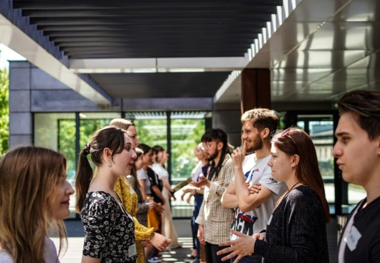 A group of people, slightly uncomfortable, stand in two lines facing each other under a modern outdoor structure, deeply engaged in talk.