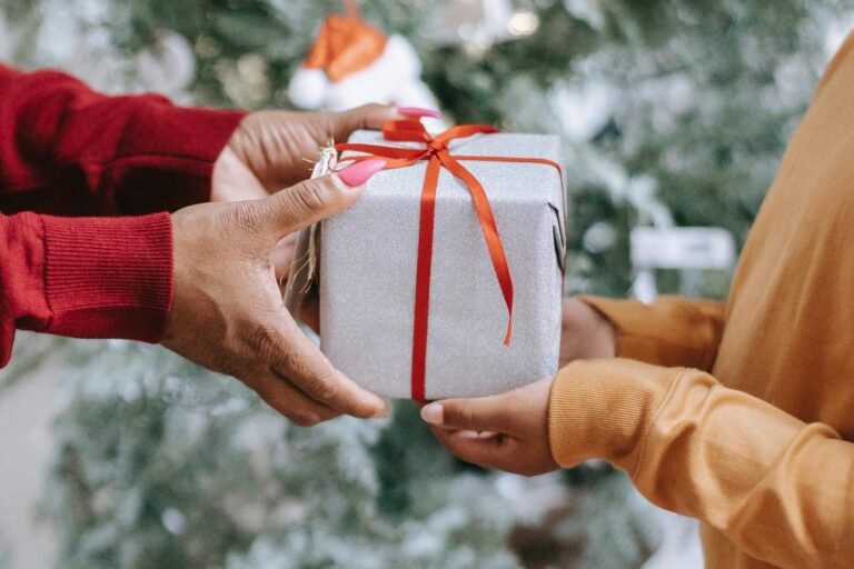 Amidst the soft glow of a blurred Christmas tree, two people share in the joyful ritual of gift-giving, exchanging a meaningful silver gift box adorned with a vibrant red ribbon.