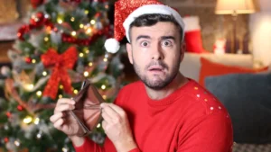 A man in a Santa hat and red sweater holds an empty wallet, looking surprised, pondering what to do now. A decorated Christmas tree glows warmly in the background, reminding him of the holidays and how he may have overspent.