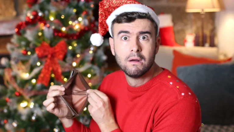 A man in a Santa hat and red sweater holds an empty wallet, looking surprised, pondering what to do now. A decorated Christmas tree glows warmly in the background, reminding him of the holidays and how he may have overspent.