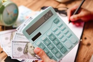 A person focuses on simple money management, holding a calculator displaying 207.25 and jotting notes beside a stack of hundred-dollar bills. In the background, an alarm clock softly ticks towards 2025, a subtle reminder of time's passage.