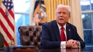 A man in a suit sits at a desk in an office with flags and large windows in the background, contemplating the impact of Trump Tariffs on consumers as 2025 approaches.
