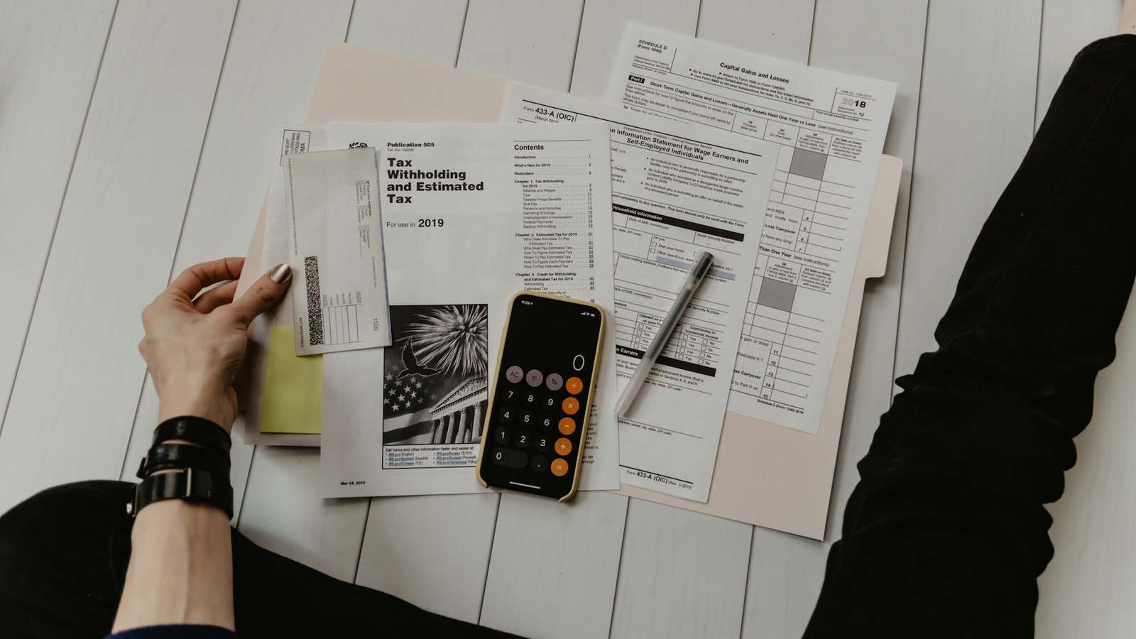 Person sitting on floor with tax documents, a pen, check, smartphone with calculator app open, and a folder, carefully preparing financial paperwork as part of their debt management plan. Only arms and legs visible.