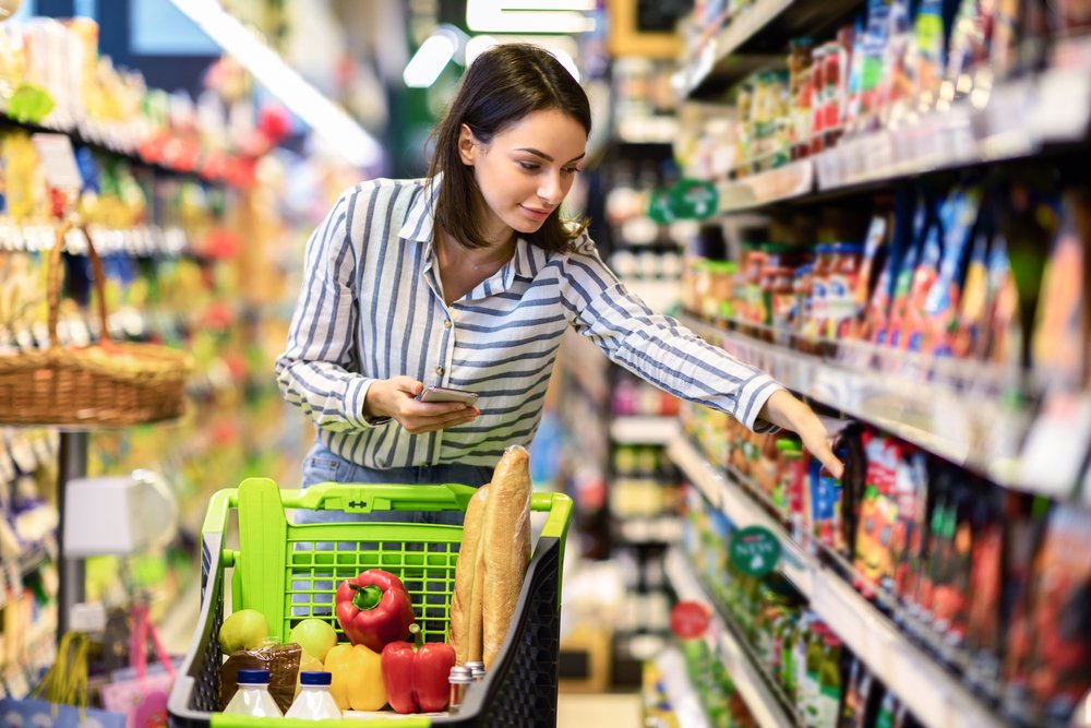 A woman pushes a shopping cart with groceries and reaches for an item on a grocery store shelf while holding her phone, using smart budget hacks to help save money on her grocery budget.