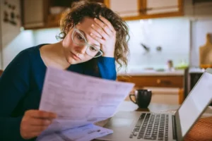 A person wearing glasses looks stressed while reviewing their credit report at a kitchen table, with a laptop and coffee mug nearby.