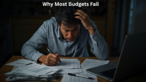 A man sits at a table with a laptop and paperwork, appearing stressed as he works, with the text "Why Most Budgets Fail" above him—highlighting the challenges and common causes of budgeting failure.