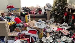 A living room filled with unwrapped presents, torn wrapping paper, toys, and boxes scattered around a Christmas tree and sofa—a reflection of holiday wants and joyful chaos.