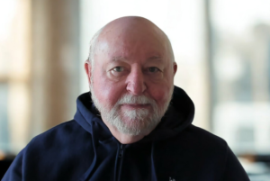 An older man with a white beard and short hair is wearing a dark hooded sweatshirt, looking directly at the camera with a neutral expression.
