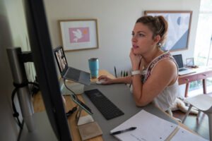 A woman stands at a desk, using a computer and laptop, with a notebook and pen beside her. She appears to be participating in a video call in a home office, possibly discussing tax preparation tips during tax season.