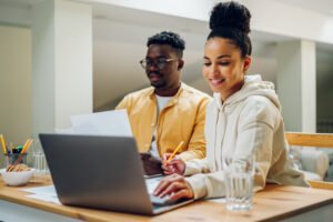 Two people sit at a table with a laptop, papers, pencils, and glasses of water, working together in a well-lit room as they conduct a simple financial checkup to set themselves up for a better year.