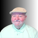 An older man with a light beard wearing a beige flat cap and a light green collared shirt, smiling in front of a black-to-white gradient background. An older man with a light beard wearing a beige flat cap and a light green collared shirt, smiling in front of a black-to-white gradient background.